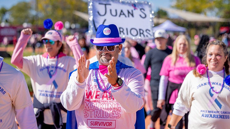 Participant in a pink breast cancer awareness walk claps while walking with a group, wearing a blue hat, cape, and “Survivor” shirt; a “Juan’s Warriors” sign is visible in the background.