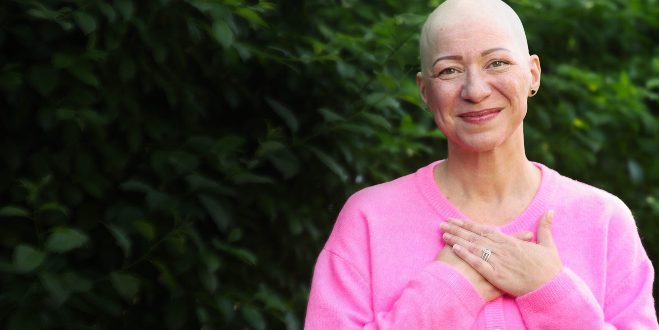 Person wearing a pink sweater, standing outdoors with hands over chest against a blurred green leafy background.