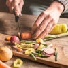 A person's hands are shown cutting a carrot with a knife on a cutting board surrounded by other vegetables.