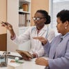 A female doctor points toward a computer screen on the desk where she sits next to a woman in a gray sweater. 