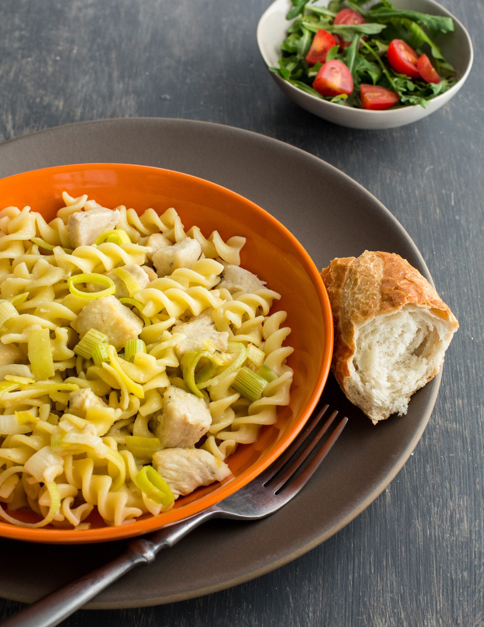 An orange bowl filled with a mix of fusilli and chicken sits on a gray plate next to a fork and a piece of a baguette.
