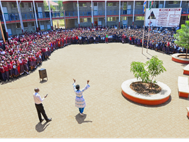 Two presenters stand in a large courtyard addressing a crowd of people lined up around the perimeter