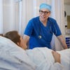 A doctor wearing blue scrubs and glasses talks to a woman in a hospital bed.