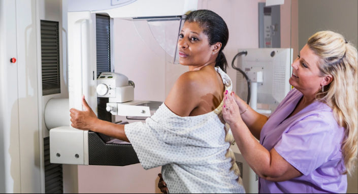 A dark-haired woman wearing a hospital gown gets a mammogram with assistance from a blonde woman wearing light purple scrubs.