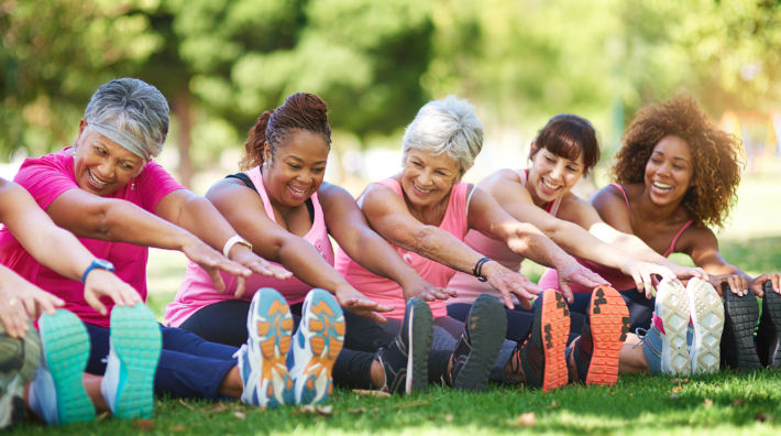 Five women wearing workout clothes sit on the ground and stretch their arms toward their toes. 