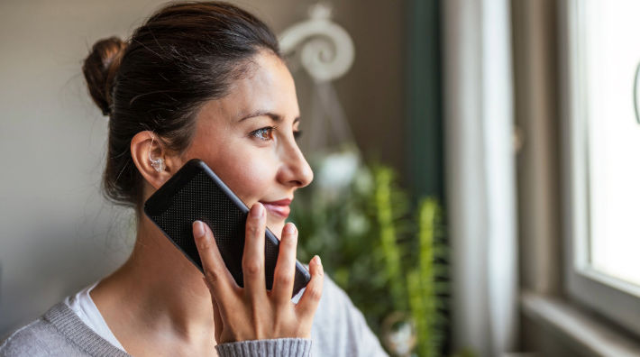A woman with a hearing aid holds a smartphone next to her face as she looks out a window.