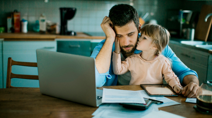 young daughter kisses her stressed out dad while he works on laptop at kitchen table