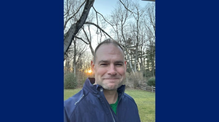 Andy Whitehouse stands in front of trees as the sun peeks through them in the background.