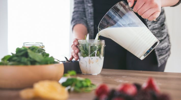 A person pours milk from a pitcher into the cup of a small blender sitting on a counter next to bowls of greens and fruit. 