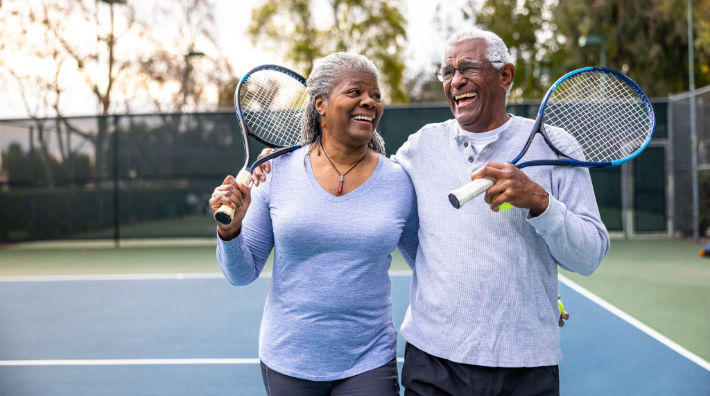 A woman and a man holding tennis rackets laugh as they walk with their arms around each other on a tennis court. 