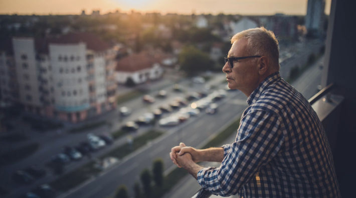 A man with gray hair stands on a balcony overlooking a street and parked cars with a city skyline visible in the distance.