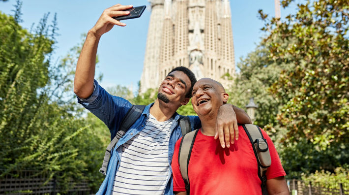 A young man holds a smartphone up to take a picture of himself and an older man in front of a building in the background.