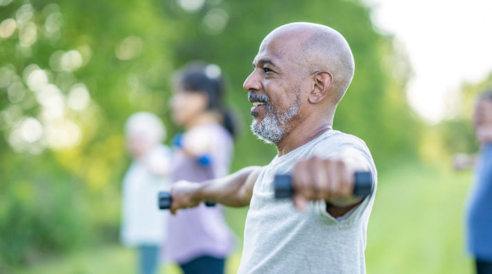 A bald man with a beard stands outdoors with arms extended and holds small weights in each hand. 
