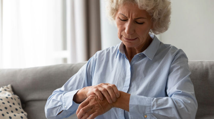 A gray-haired woman wearing a long-sleeved button-down shirt looks down as she holds her wrist with her other hand.