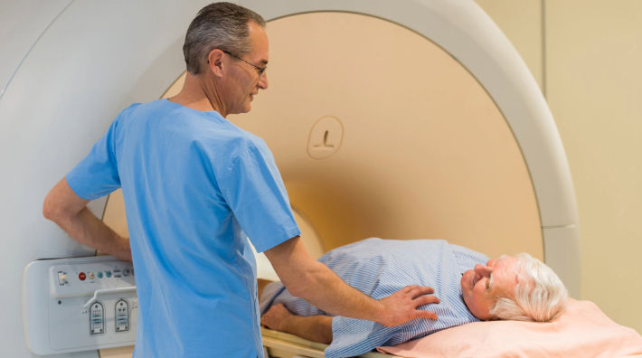 A man in blue scrubs rests his hand on the shoulder of an older woman lying on the patient table of an MRI machine.