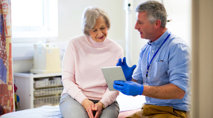 An older woman at a doctor’s office sits on an exam table and looks at a tablet held by a man in a blue shirt and gloves.