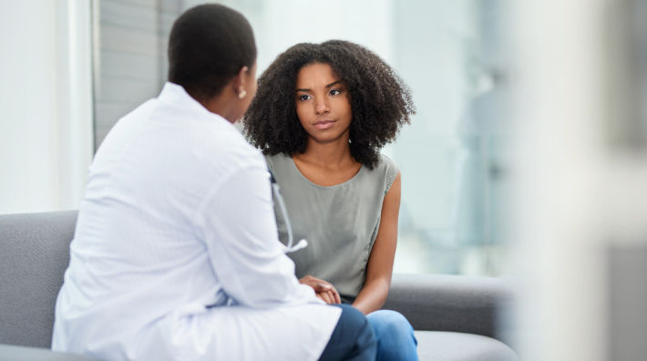 A doctor wearing a white lab coat sitting on a couch turns toward a woman wearing a gray tank top and jeans.