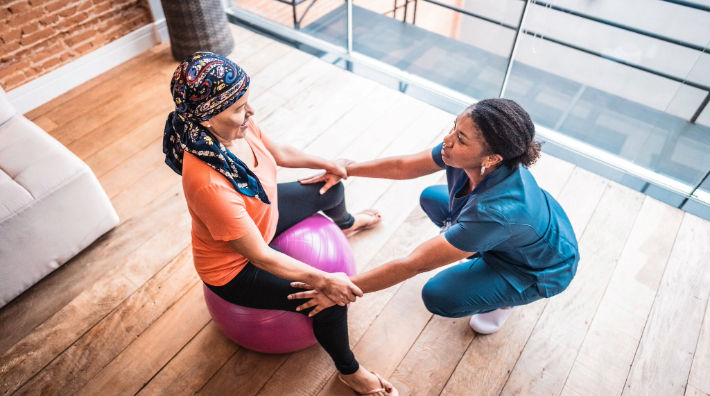 A woman, wearing a scarf on her head, sits on an exercise ball and grasps the wrists of a woman wearing scrubs who squats in front of her.