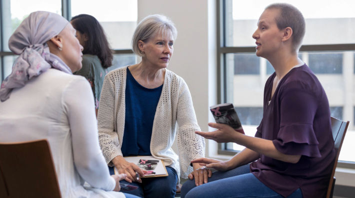 A woman in a purple blouse speaks to a woman in a headscarf and a woman in a white cardigan who both sit next to her. 