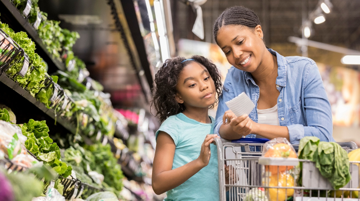 a mom and young daughter go over their shopping list in a grocery store produce aisle