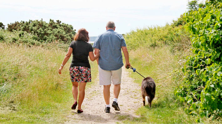 Seen from the back, a woman and a man walk a dog on a sandy seaside path with grasses and other plants on both sides.
