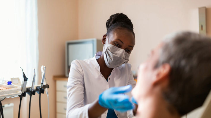 A female dentist wearing a mask extends a gloved hand toward the face of a man in a dental chair. 