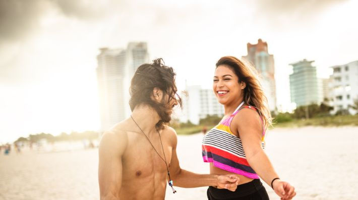 A man smiles at a woman in a striped tank top who stands beside him on a beach with buildings in the background. 
