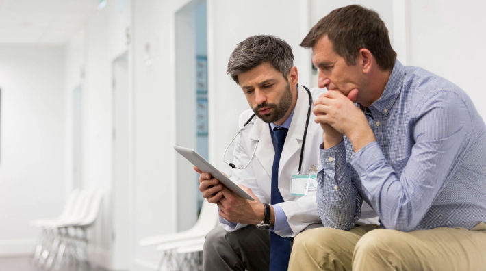 A man in a blue button-down shirt sits next to a male doctor wearing a white lab coat as they look at a tablet. 