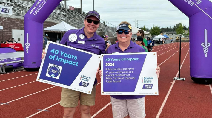 Suzy Lawrence (Left) at a Relay for Life event standing in front of a purple inflatable arch on a track