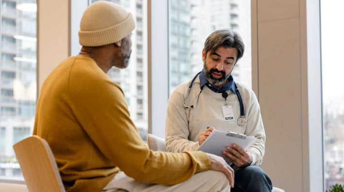 A man in a yellow sweatshirt and knit cap sits facing a bearded doctor taking notes on a clipboard. 