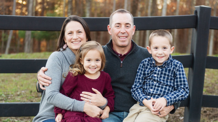 Josh Herting smiles for a photo with his wife and kids in front of a farm fence and field