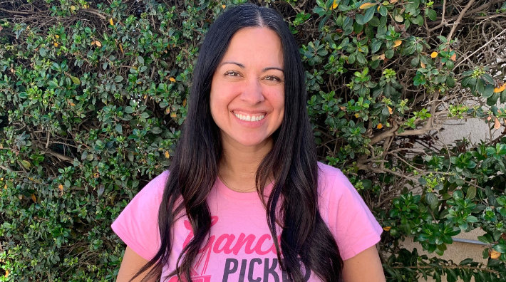 Breast cancer survivor, Drea Sauceda, smiles in pink shirt and stands in front of greenery