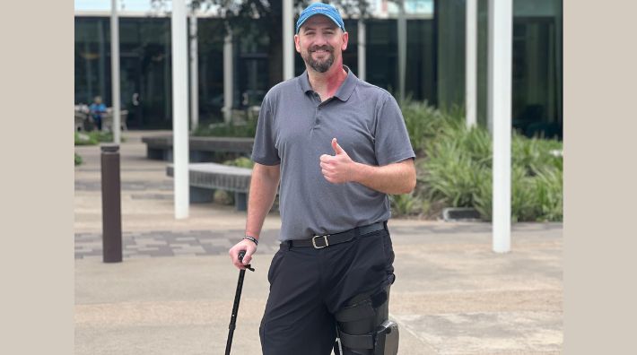 Neil Erickson smiles outside a Hope Lodge, standing with his cane and giving a thumbs‑up