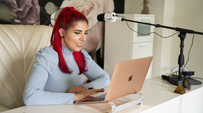 Danielle Massi, a woman with red hair and a blue shirt, smiles while typing on her rose gold laptop