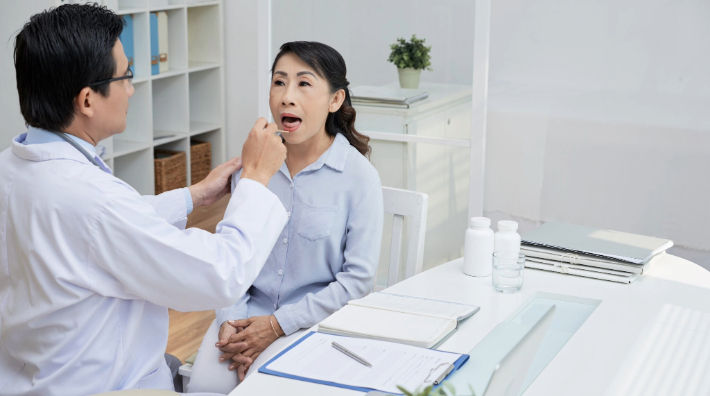 A male doctor wearing a white lab coat examines the mouth of a woman sitting in front of him. 