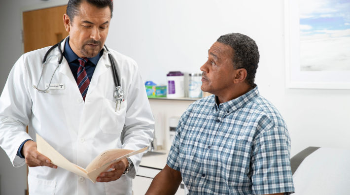 A man sitting in an exam room looks at a male doctor in a white lab coat who holds an open file folder. 