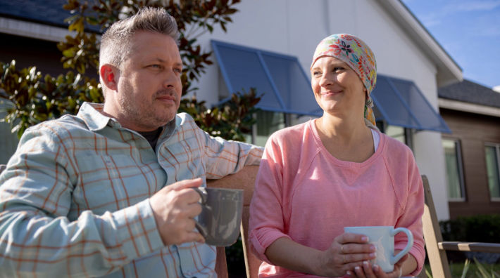A woman wearing a floral headscarf smiles at the man next to her as they sit outside holding coffee cups.