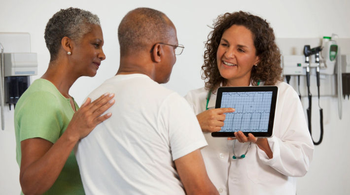 A woman rests her hand on the back of man as he looks at the screen of a tablet held by a female physician.