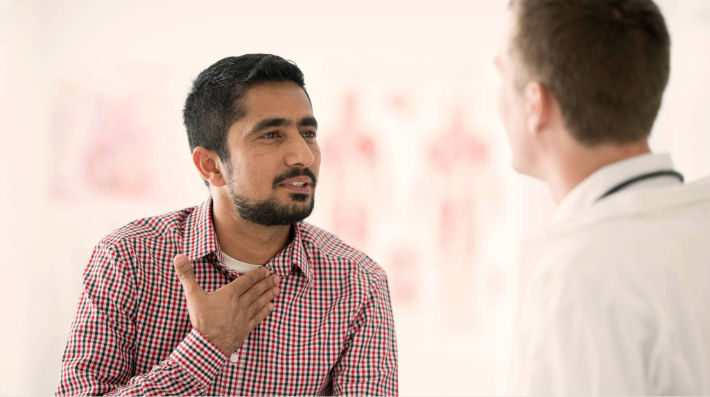 A bearded man rests his hand on the top of his button-down shirt as he faces a male doctor in a white coat, who is seen from the side. 