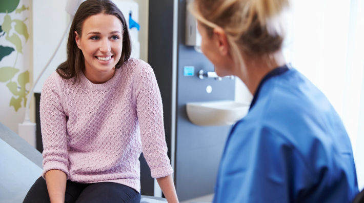 A smiling woman in a pink sweater sits on an exam table facing a woman in blue scrubs with a sink in the background. 