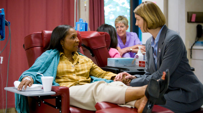 A woman in a blazer places her hands on the arm of a womanreceiving a chemotherapy treatment as she sits in a recliner. 