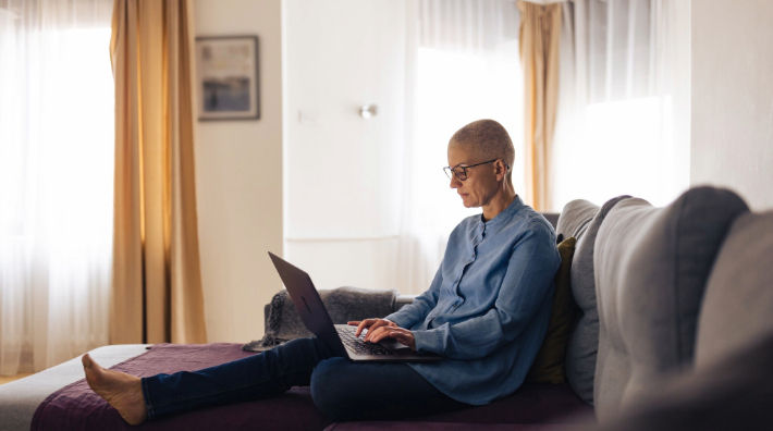 A bald person wearing a blue top and dark pants sits on a bed with one leg outstretched as they type on a laptop. 