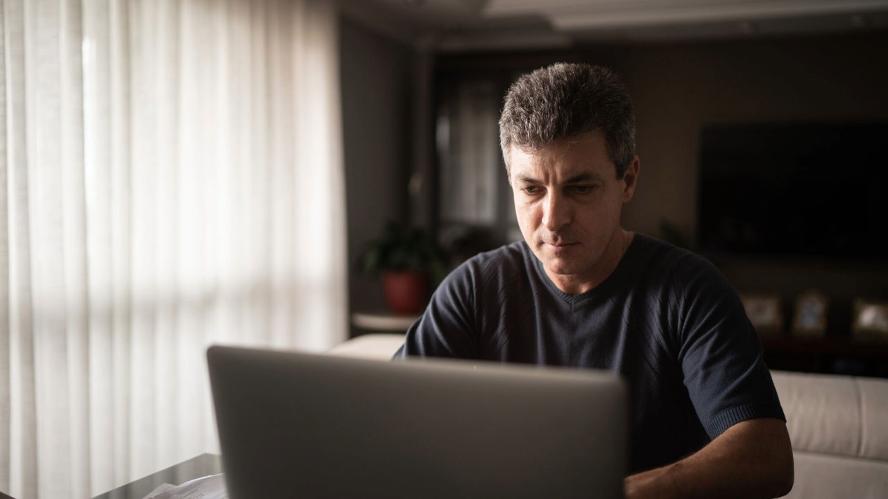 A man wearing a black T-shirt sits and looks at a laptop computer in a shadowy room with a window with off-white curtains.