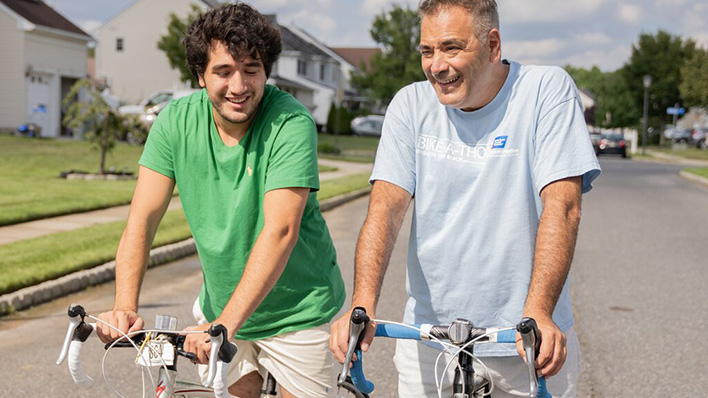 Two men smiling while riding bicycles on a suburban street.