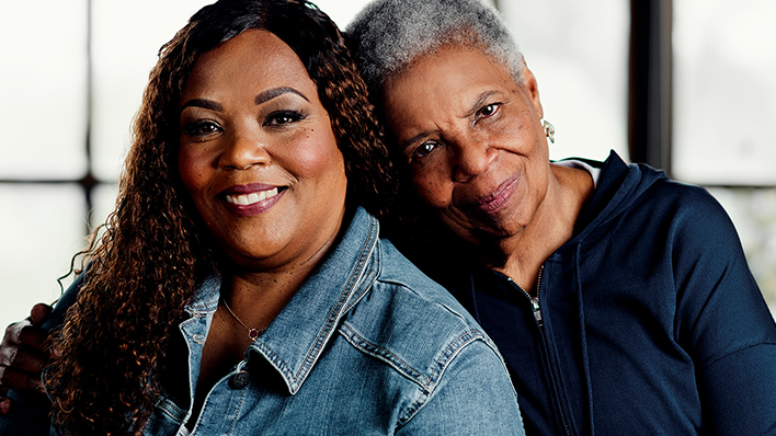 Two people smiling at the camera, one leaning on the other's shoulder, with a blurred background."