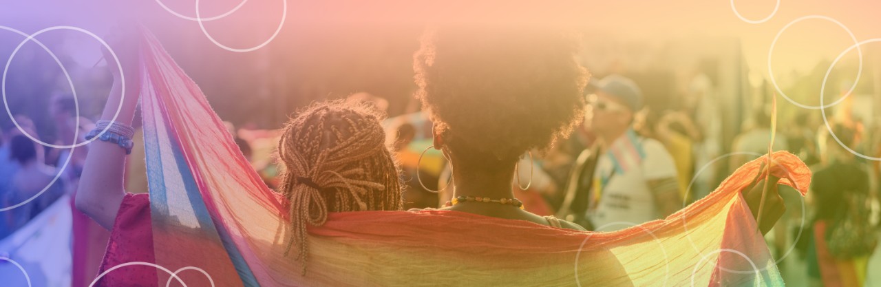 two women at an LGBTQ+ event with flag draped around their backs