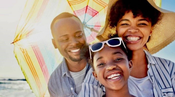 close up of smiling mom, dad and young daughter under umbrella at the beach with sun shining in background