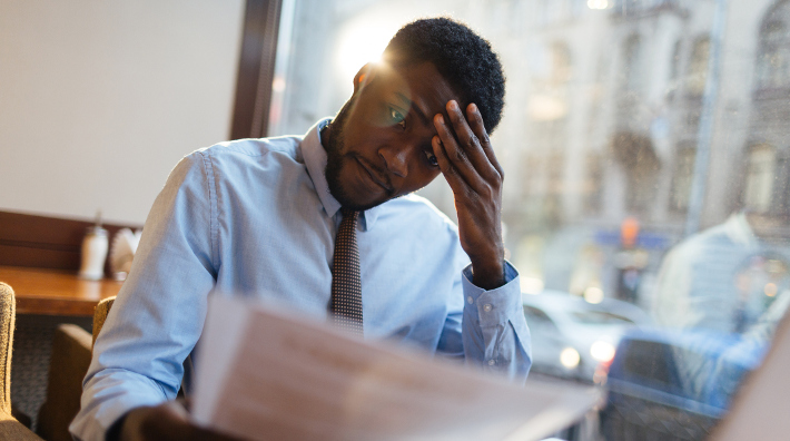 a stressed out man rubs his brow as he looks at papers while sitting at a computer