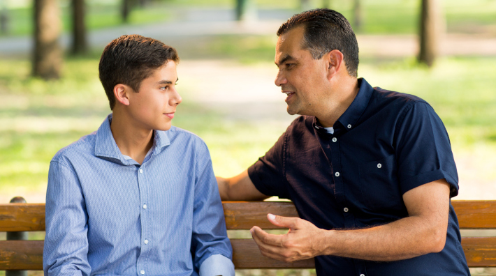 Father and his teenage son sitting on a bench, talking and looking at each other  