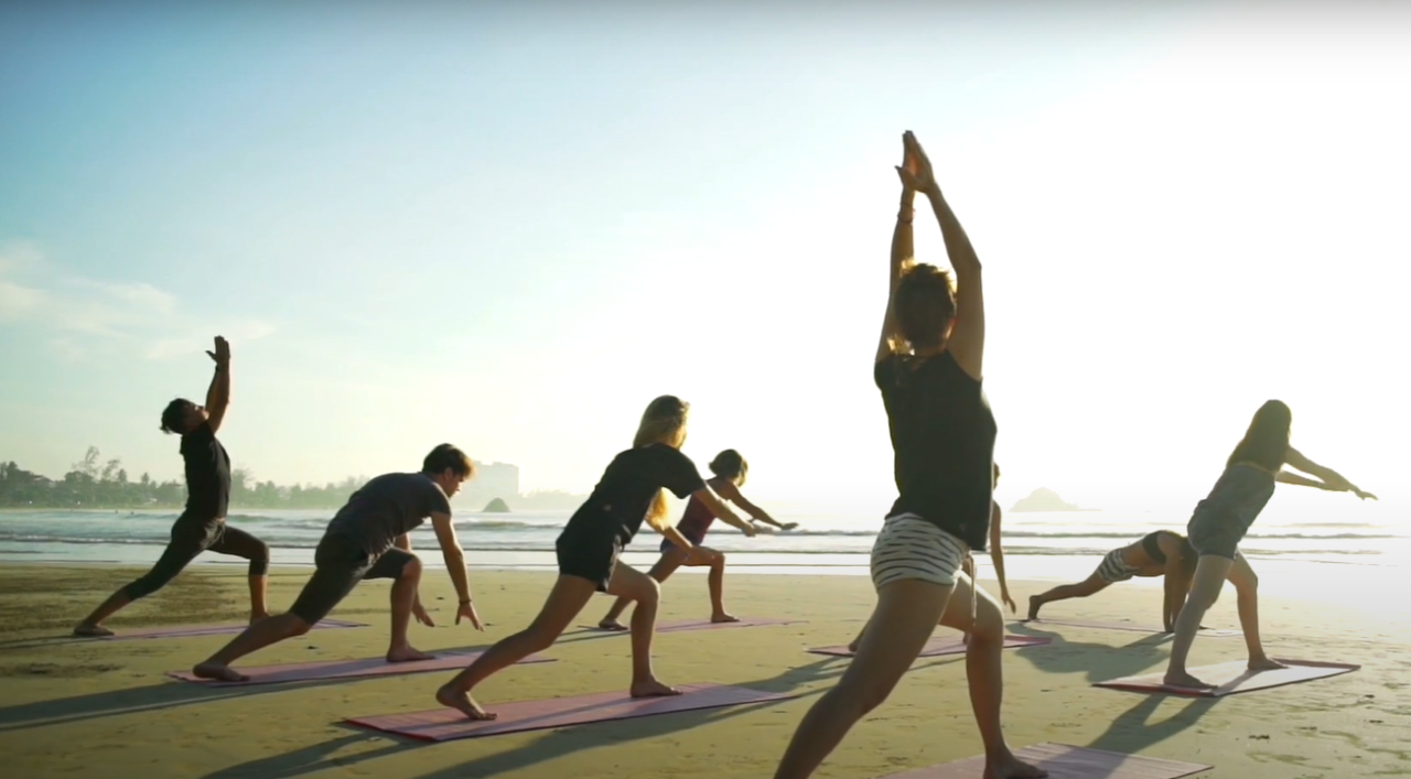 Imagen estática del video mostrando un acercamiento de un grupo de personas practicando yoga en la playa.